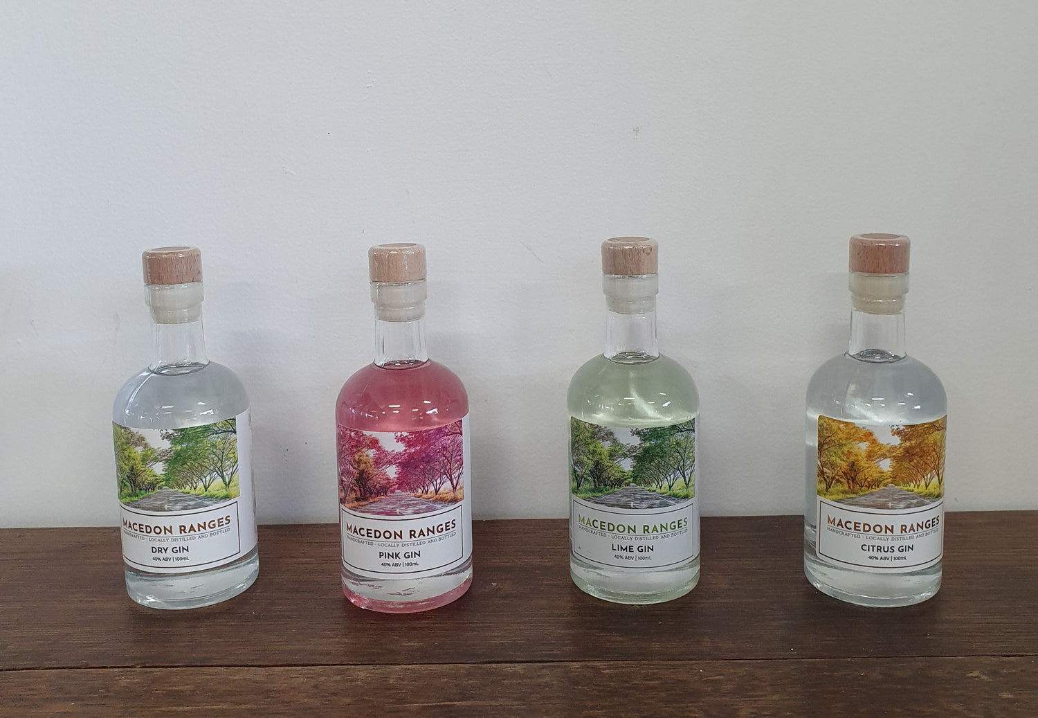 Four bottles of Macedon Ranges Gin in different flavors, including Pink Gin, Lime Gin, Citrus Gin, and Dry Gin, displayed on a wooden surface against a white background.