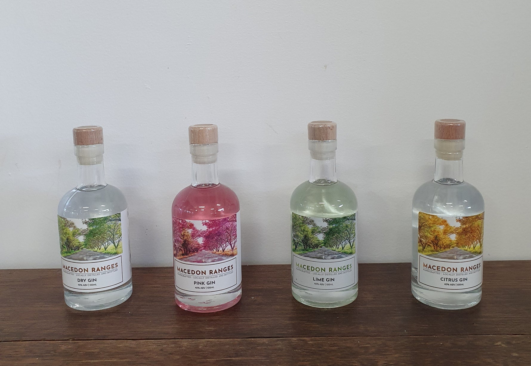 Four bottles of Macedon Ranges Gin in different flavors, including Pink Gin, Lime Gin, Citrus Gin, and Dry Gin, displayed on a wooden surface against a white background.
